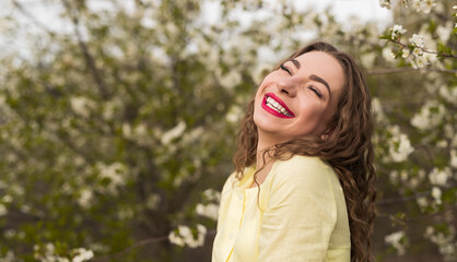 A joyful young woman wearing a yellow top, smiling brightly while surrounded by blooming trees in a spring garden.