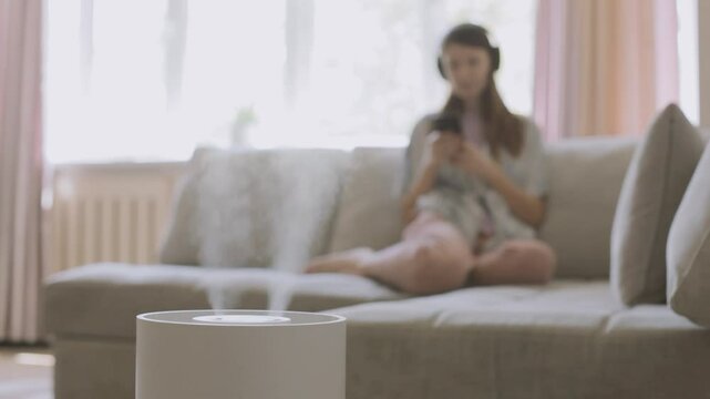 Air humidifier spraying fresh water while woman sitting on sofa and listening music. 