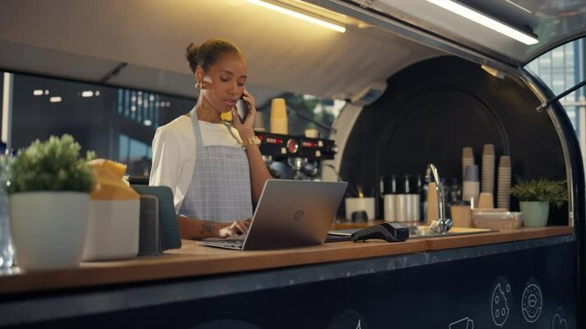 African American Female Food Truck Owner Multitasks, Using a Laptop Computer and Talking on a Phone. Small Business Manager Working Inside a Food Stand with Cups, Plants and Coffee Machines