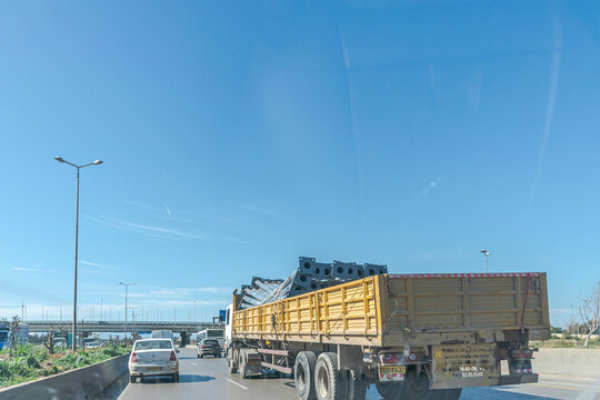 Blida, Algeria - February 16, 2022: A truck carrying metal poles loaded on the back. Driving near cars in second position on the multilane highway traffic. Sunny day and blue clear sky. 