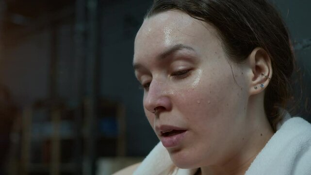 Zoom in shot of exhausted young sportswoman wiping sweat from her face with hand and towel while sitting on floor in gym after intense workout