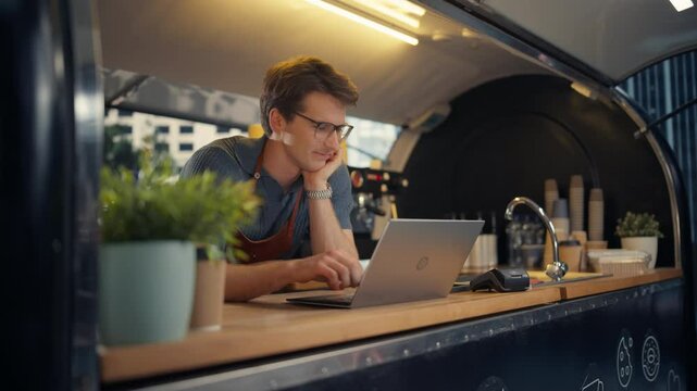Handsome Male Street Cafe Staff Member Working in a Food and Drinks Truck. Young Caucasian Man Managing Online Orders on a Laptop Computer, Ordering Supplies of Coffee and Tasty Snacks