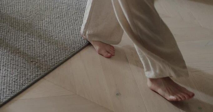 Close-up of bare feet leisurely walking indoors on a wooden floor and transitioning to a carpeted area, conveying a sense of comfort and relaxation. Bare Feet Walking Indoors On Wooden Floor