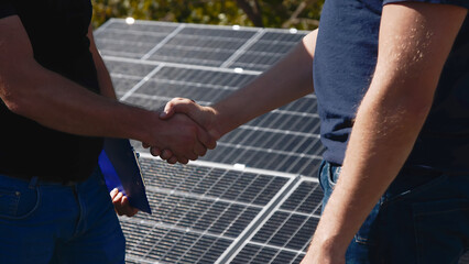 Close-up of two engineers shaking hands with solar panels in the background. Highlights a...