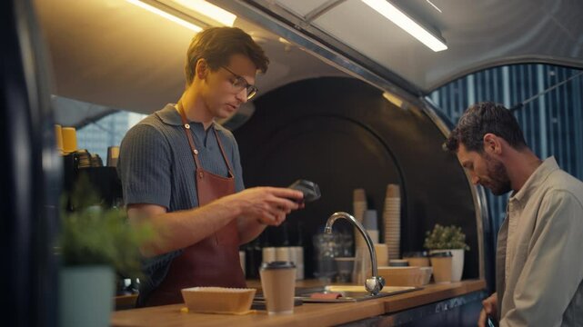 Caucasian Customer Buying Coffee and Snacks To Go From a Trendy Street Cafe in a Modern City Neighbourhood. Barista Serves Order with a Smile, Client Makes a Transaction with His Smartphone