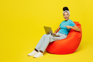 Full body young Latin IT woman wears blue top t-shirt headband casual clothes sit in bag chair use work point index finger on laptop pc computer isolated on plain yellow background. Lifestyle concept.