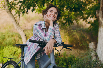 Young happy pensive woman she wearing checkered shirt casual clothes look camera walking stand lean on bicycle rest relax in spring sunshine green city park outdoors on nature. Urban leisure concept.