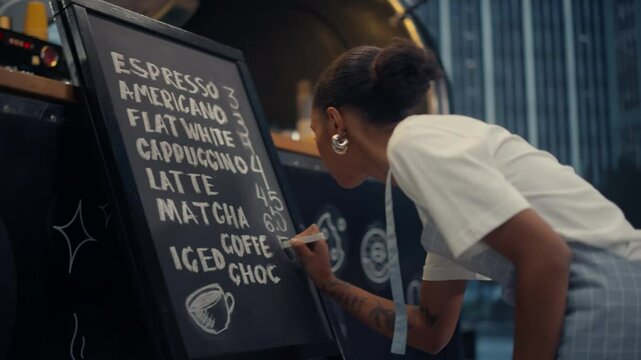 African American Young Woman Writing a Drinks Menu on a Chalkboard Outside a Street Cafe, Listing Various Coffee Drinks: Espresso, Americano, Flat White, Cappuccino, Latte, and Matcha