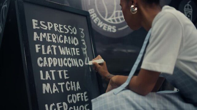 Black Female Food Truck Worker in an Apron Writes a Coffee Menu on a Chalkboard, Listing Drinks Like Espresso, Cappuccino, Latte, Matcha. Writing Down Prices and Preparing to Welcome Clients
