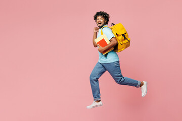 Full body side view young happy Indian boy student wear blue t-shirt casual clothes backpack bag hold books jump high run fast isolated on plain pink background High school university college concept
