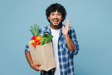 Young Indian man in shirt hold brown craft bag for takeaway mock up with food products scream share hot news about sales hands near mouth isolated on plain blue background Delivery service from shop