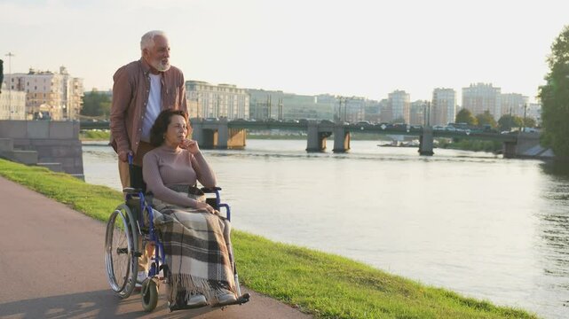 Senior woman in wheelchair walking with caregiver old man on road in park. Elderly family couple man husband supporting embracing woman wife in chair for people with disability outdoor. Rehabilitation