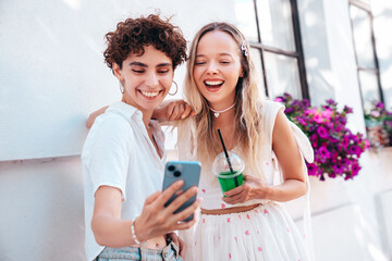 Two young beautiful smiling hipster female in trendy summer clothes. Carefree women posing outdoors. Positive models holding cocktail smoothie drink in plastic cup, use mobile smartphone apps