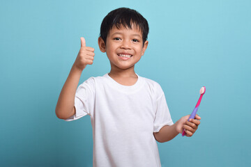 A happy Asian boy holding a toothbrush and giving a thumbs up isolated on blue background. Boy smiles showing his teeth. Children's dental care and hygiene