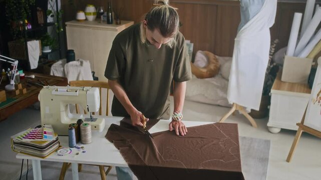 View from above of Caucasian gen Z designer making cuts on piece of fabric while tailoring at studio