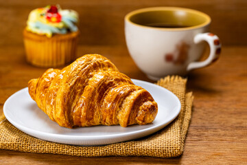 Freshly baked delicious french croissant.. Side view of Freshly baked delicious french croissant in white ceramic dish on sack cloth with a cup of black coffee and cupcake on wooden table.