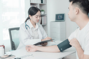 Fototapeta premium Asian female nurse talking and asking about symptoms of young male patient using blood pressure monitor to measure blood pressure level Medical examination result during consultation