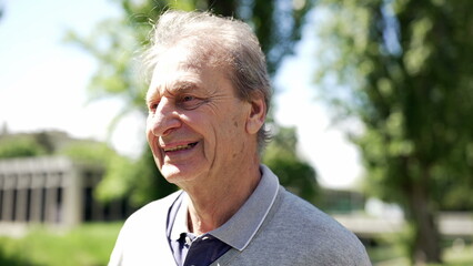 Elderly man smiling outdoors, close-up, enjoying sunny day in park, surrounded by trees, happy and content expression, senior lifestyle, casual attire, natural background, golden years joy