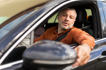 Serious middle aged businessman touching and looking at side mirror during car test-drive, before choosing buying new auto