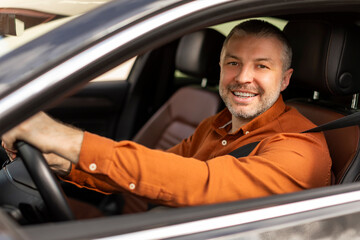 Positive middle aged businessman driving his luxury auto, looking through open window and smiling,...