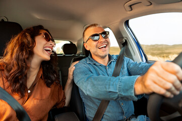 Excited playful European couple in sunglasses driving luxury car, enjoying music, singing and smiling, spouses going on vacation, windshield view