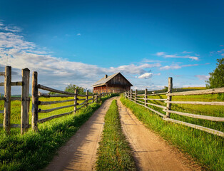 Fototapeta premium Landscape with a dirt road between wooden fences leading to an old wooden house in a field with green grass