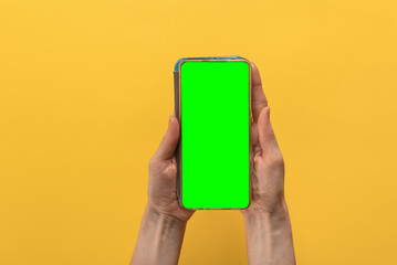 Closeup of woman holding modern smartphone with green screen for advertising on yellow background. Hand holding a smartphone with a flat chromakey screen, top view.