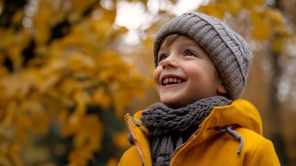 A young boy, clad in a yellow coat, gray hat, and scarf, revels in the vibrant autumn foliage of a park, his cheerful countenance radiating joy.