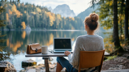 Fototapeta premium A woman working remotely with a laptop by a calm mountain lake, surrounded by nature's beauty during autumn.