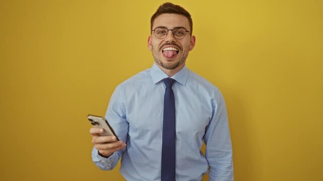Young hispanic man wearing shirt and tie sticking tongue out happily while using smartphone over isolated yellow background - funny and expressions of joy and fun!
