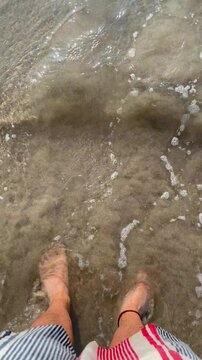 Vertical video of a man's feet on beach vacation on the shore and crystal clear water