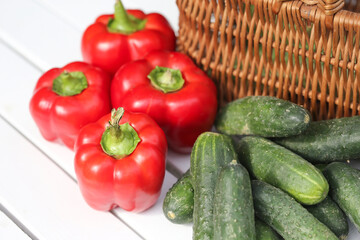 Fresh red bell peppers. Green cucumbers on a white wooden table. Vibrant vegetables. Variety of colors and textures. Healthy produce perfect for salads and cooking. Wicker basket of vegetables.