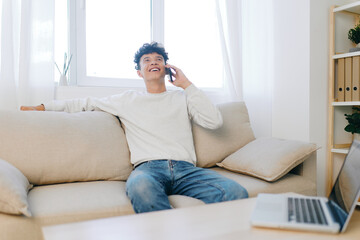 Fototapeta premium Young man talking on the phone while sitting on a comfortable couch in a bright, modern living room with a natural light background Relaxed and cheerful atmosphere, cozy design