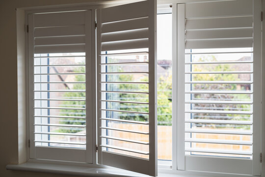 Three frames of  white modern wooden shutters across a window. One section is open. The shutter slats are swivelled in different positions.
