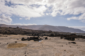 White sand beach with black rocks of volcanic origin. Mountains in the background and sky with big white clouds. Caletón Blanco, Lanzarote, Canary Islands, Spain