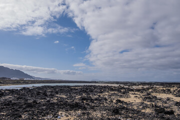 White sand beach with black rocks of volcanic origin. Mountains in the background and sky with big white clouds. Caletón Blanco, Lanzarote, Canary Islands, Spain