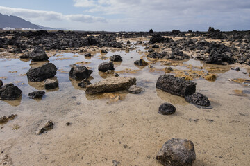 White sand beach with black rocks of volcanic origin. Mountains in the background and sky with big white clouds. Caletón Blanco, Lanzarote, Canary Islands, Spain