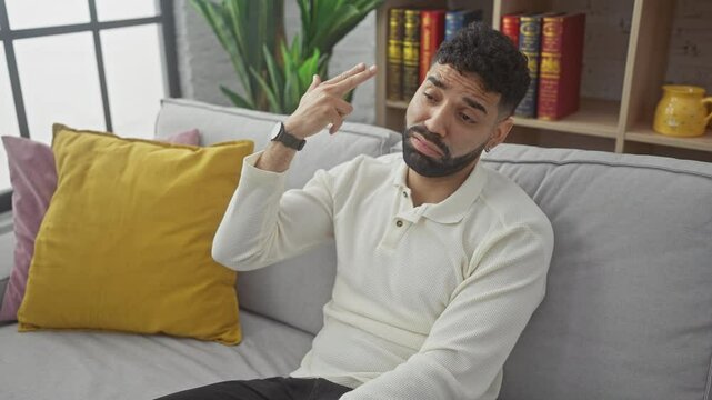 Young hispanic man at home, making a suicide gesture by pointing fingers like a gun towards his head, shooting imaginary bullet as symbol of stress, depression and panic