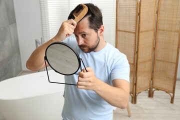 Man brushing his hair near mirror indoors. Alopecia problem