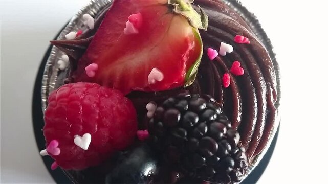Top down view of chocolate mud cake with heartshaped sprinkles, strawberry, raspberry and blackberry topping in foil cupcake liner.