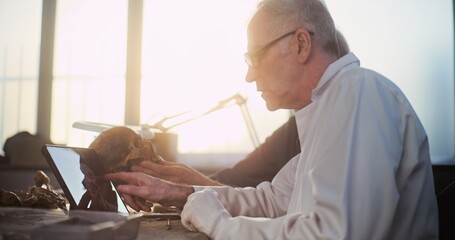 Archaeological lab: Two researchers or scientists examine fossil skull, have discussion, study 3D model and visualization of prehistoric extinct human head displayed on digital tablet computer screen.