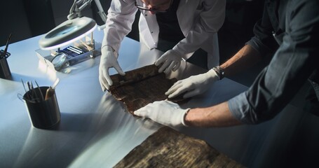 Two archaeologists in gloves carefully roll out ancient Egyptian papyrus on the table in...