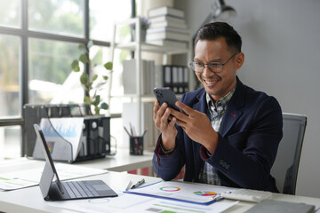 Cheerful businessman is checking his smartphone while sitting at his desk in a bright office