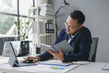 Fototapeta premium Asian accountant is working in his office, checking some numbers on a digital tablet and reading a book