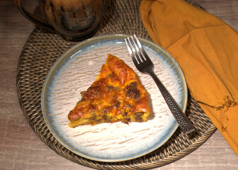 Top down photo of a sausage quiche on a ceramic plate with a fork, napkin, and mug.  The quiche is a single slice. 