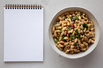 Homemade White Bean Salad with Herbs and Onion in a Bowl, blank notepad, top view. Flat lay, overhead, from above.