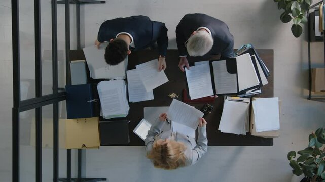 Top down view of two employees standing in front of boss at office desk showing documents to her