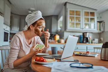 Woman with face mask enjoying breakfast and coffee while working on laptop in kitchen