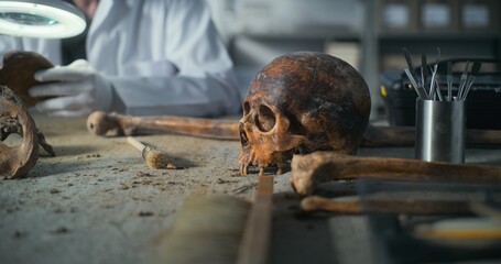 Archaeological laboratory: Fossil skull of extinct prehistoric man and professional tools on the table. In the background, archaeologist or scientist works with bones remains using magnifying lamp.