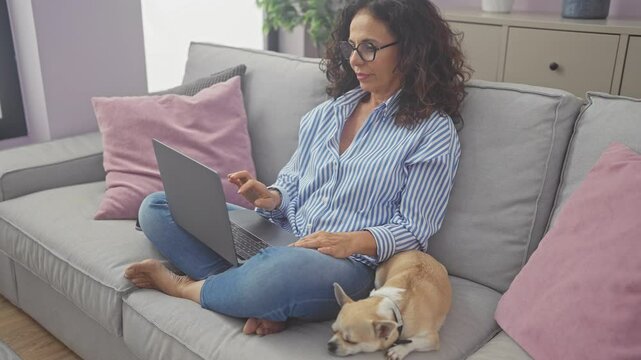 Middle-aged woman with glasses using laptop next to sleeping chihuahua on couch indoors